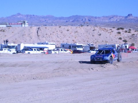 1990 Ford Bronco - blue Red Bull-liveried off-road race truck drives a sandy desert course, RVs and a casino on the bluff behind.