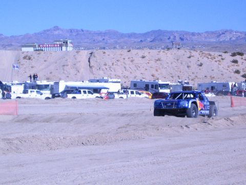 1990 Ford Bronco - blue Red Bull-liveried off-road race truck corners on a sandy desert course with a hilltop casino in the background.