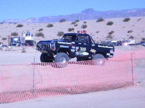 1990 Ford Bronco - black number 300 off-road race truck catches air over a sandy desert course behind orange safety fencing.