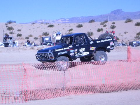 1990 Ford Bronco - black number 300 off-road truck, covered in sponsor decals, runs a desert race course behind orange fencing.