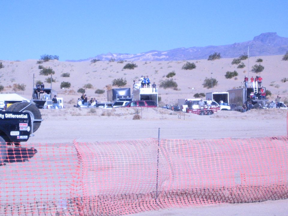 Desert off-road race staging area behind orange safety fence with spectators on platforms