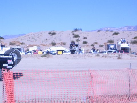 1990 Ford Bronco - Trailers and crews gather in a sandy desert race pit area, seen past an orange safety fence under a clear blue sky.