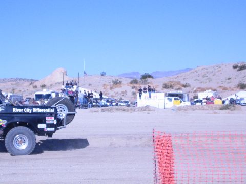1990 Ford Bronco - black off-road buggy with a River City Differential spare-tire cover sits at a sandy desert race pit with crews behind it.