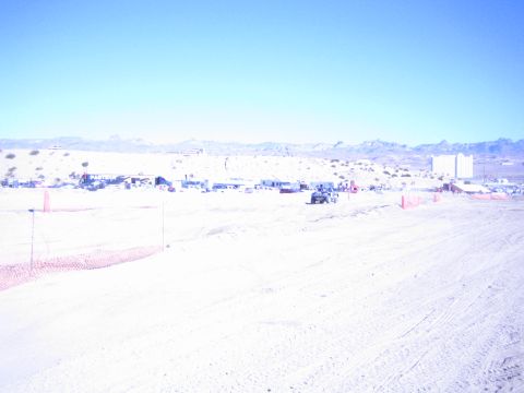1990 Ford Bronco - bright, washed-out wide shot of a sandy desert race course lined with orange fencing and distant pit structures.