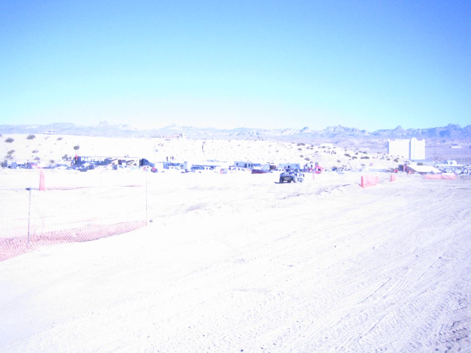 Wide bleached desert race course with orange fencing and distant pits under blue sky