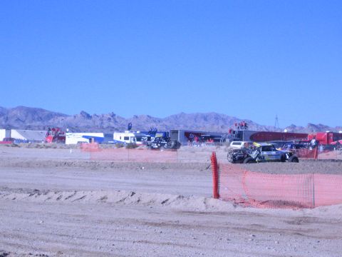 1990 Ford Bronco - Trailers and an off-road race truck line a desert pit area beyond a dirt berm and orange fence, with mountains behind.