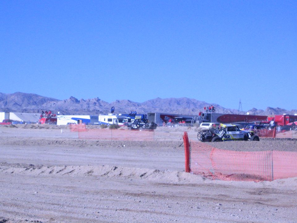 Desert race pits with trailers and an off-road race truck behind orange fence and dirt berm