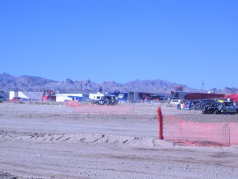 1990 Ford Bronco - row of trailers and race vehicles fills a desert pit beyond a dirt berm and orange safety fence under blue sky.