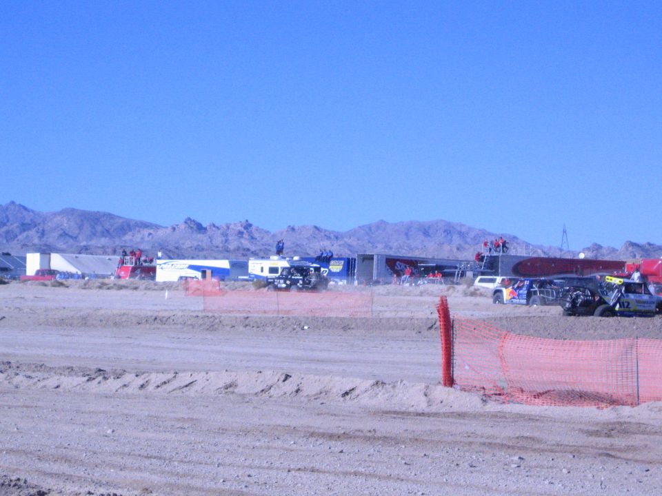 Desert race pit row with trailers and race trucks beyond a dirt berm and orange fence