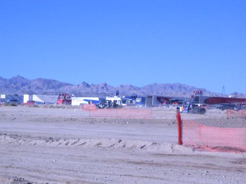 1990 Ford Bronco - Trailers and race vehicles sit across a desert pit area behind a sandy berm and orange fence, with mountains beyond.