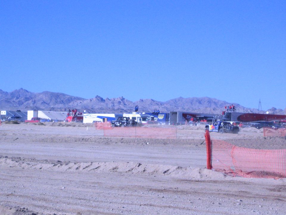 Desert race pit area with trailers and vehicles beyond a sandy berm and orange fence
