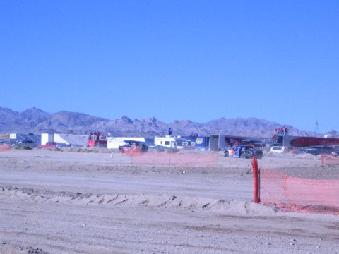 1990 Ford Bronco - spread of trailers and race vehicles, including an orange one, sits across a desert pit beyond a dirt berm and fence.