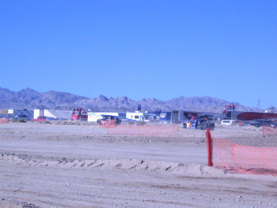 Desert race pits with trailers and an orange vehicle behind a dirt berm and orange fence