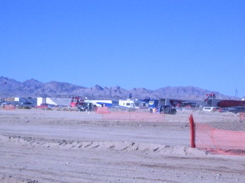 1990 Ford Bronco - distant row of trailers and race vehicles lines a desert pit area behind orange fencing and a sandy berm.