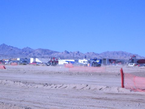 1990 Ford Bronco - Trailers and vehicles raise faint dust across a desert pit area beyond an orange fence and dirt berm, mountains behind.