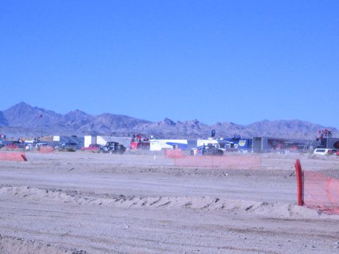 1990 Ford Bronco - long row of trailers and race vehicles sits across a desert pit beyond a dirt berm and orange fence under blue sky.