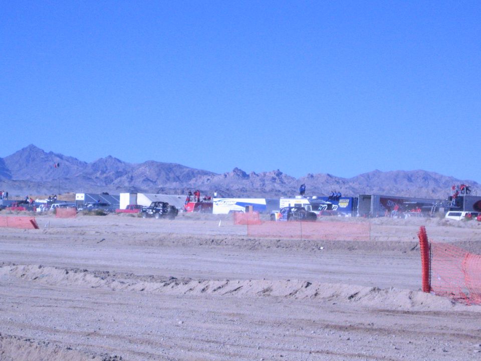 Distant desert race pit row of trailers and vehicles behind orange fence and berm