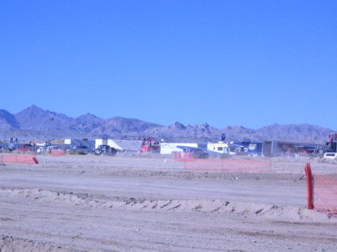 1990 Ford Bronco - Trailers and race vehicles line a desert pit area beyond orange fencing and a dirt berm, with mountains in the distance.