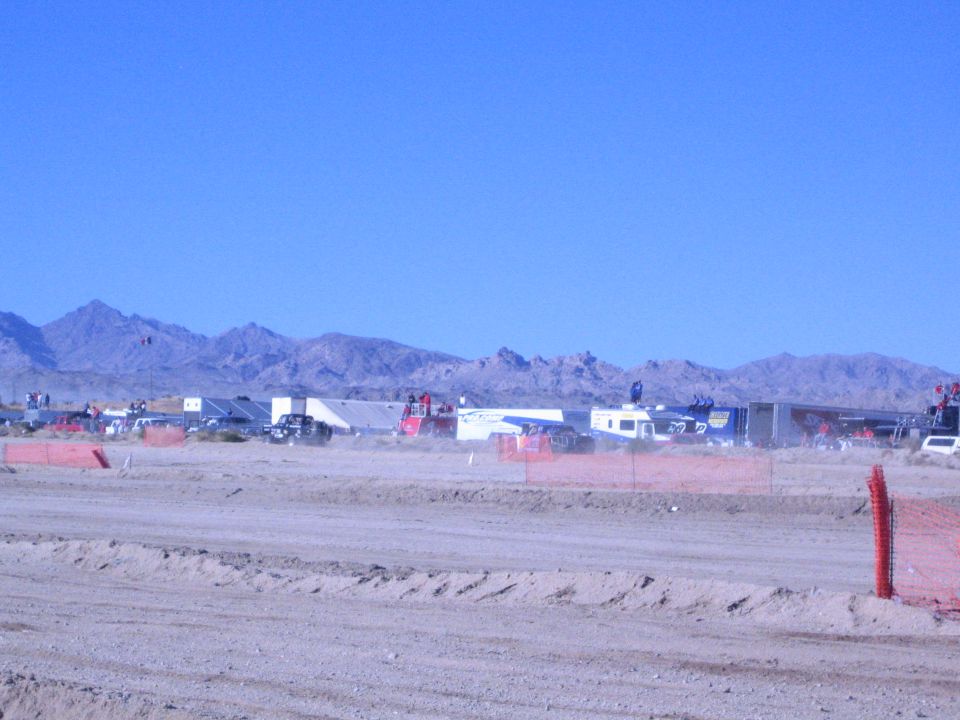 Desert race pits with trailers and vehicles behind orange fencing and a dirt berm