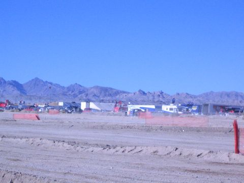 1990 Ford Bronco - distant spread of trailers and race vehicles fills a desert pit behind orange fencing and a sandy berm under blue sky.