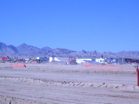 1990 Ford Bronco - Trailers and race vehicles line a wide desert pit area beyond orange fencing and a dirt berm, with mountains behind.
