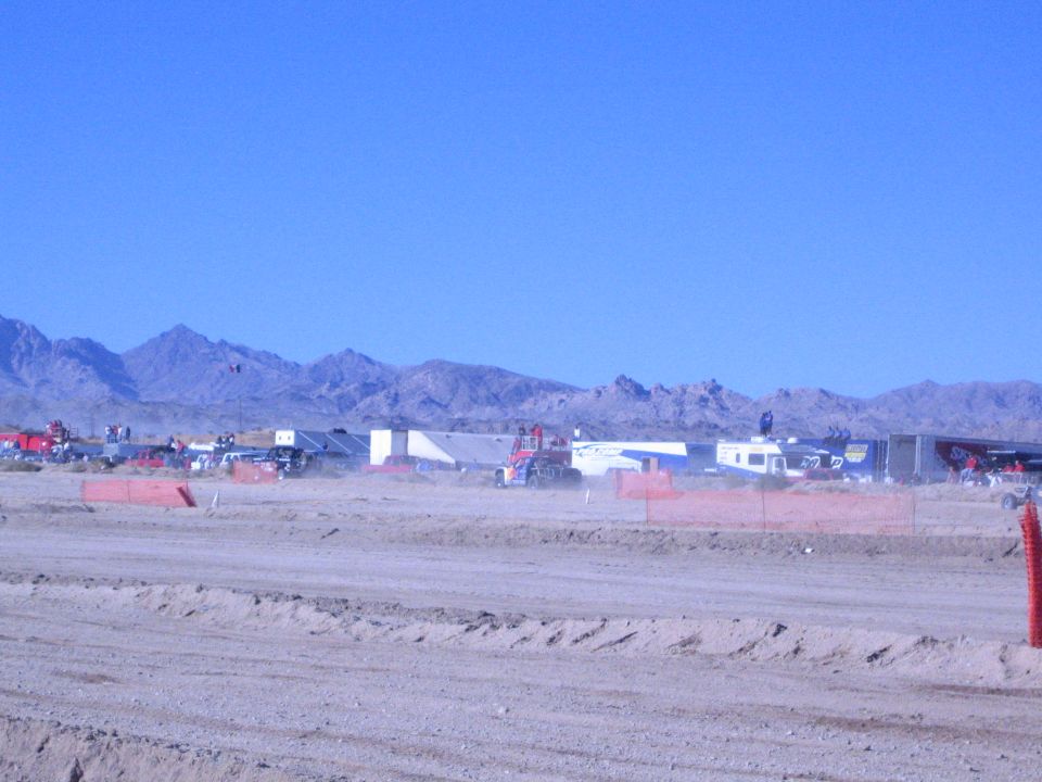 Desert race pits with trailers and vehicles behind orange fencing and dirt berm