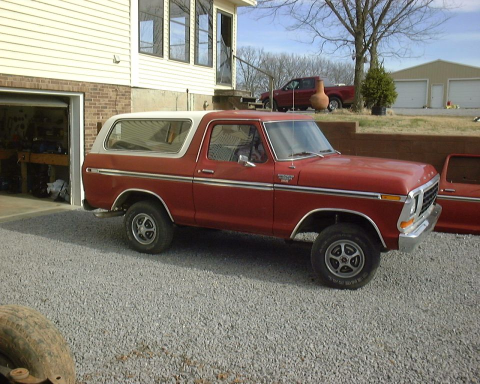 1978 Ford Bronco parked on gravel in front of a garage, another red truck in background