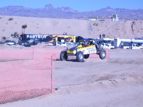 1990 Ford Bronco - yellow-and-blue off-road race buggy runs a sandy desert course past a FABTECH banner and orange fencing.