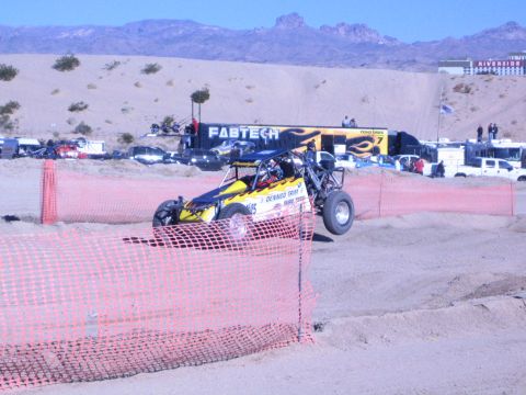 1990 Ford Bronco - yellow-and-blue off-road buggy kicks up dust on a desert course in front of a FABTECH banner and orange fencing.