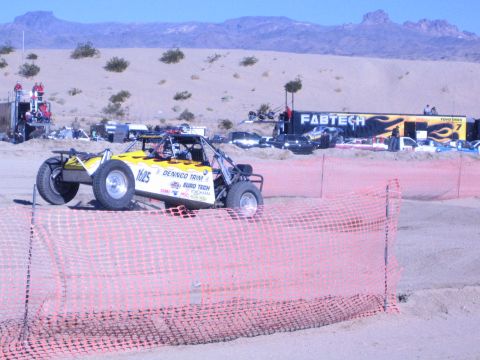 1990 Ford Bronco - yellow off-road buggy marked 1625 rounds a sandy desert course past a FABTECH banner and orange fencing.