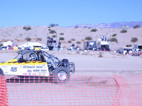 1990 Ford Bronco - yellow Dennco Trim buggy numbered 1625 runs a sandy desert course past orange fencing as spectators watch from platforms.