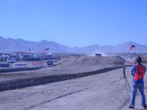 1990 Ford Bronco - photographer in a red vest with a tripod watches a distant off-road truck race past flags and banners on a desert course.