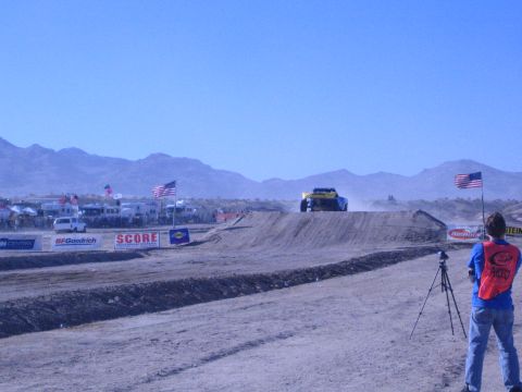 1990 Ford Bronco - yellow off-road truck crests a dirt jump while a red-vested photographer films from the foreground of a desert course.