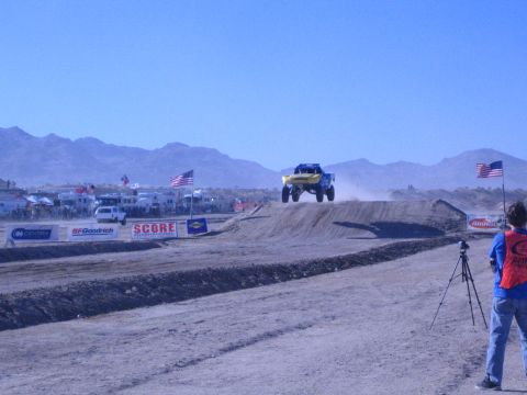 1990 Ford Bronco - yellow-and-blue off-road truck goes airborne over a dirt jump on a desert course as a photographer films nearby.