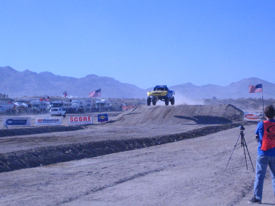 Yellow and blue off-road truck airborne over a dirt jump on a desert race course