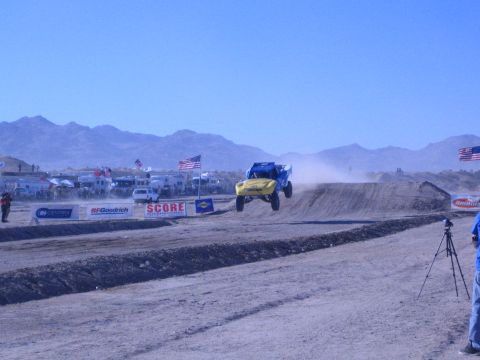 1990 Ford Bronco - blue-and-yellow off-road truck flies off a dirt jump on a desert course lined with SCORE and BFGoodrich banners.
