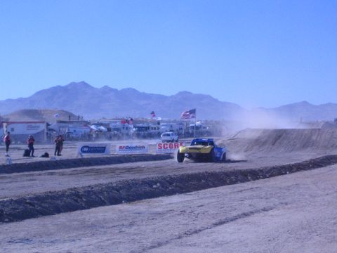 1990 Ford Bronco - yellow-and-blue off-road truck runs a sandy desert course past SCORE and BFGoodrich banners with mountains behind.