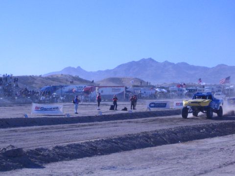 1990 Ford Bronco - yellow-and-blue off-road race truck speeds along a dirt course past BFGoodrich banners and a desert crowd under blue sky.