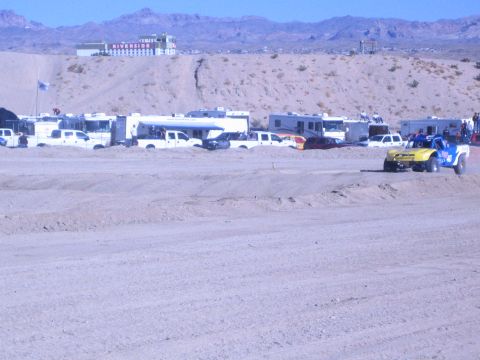 1990 Ford Bronco - yellow-and-blue off-road race truck rounds a dirt track lined with RVs and pickups below a Riverside casino on a desert hill.