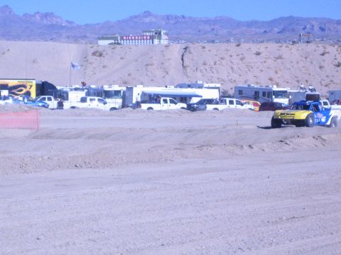 1990 Ford Bronco - yellow-and-blue race truck rolls along a graded dirt course past a row of RVs and a Toyo Tires hauler in the desert.