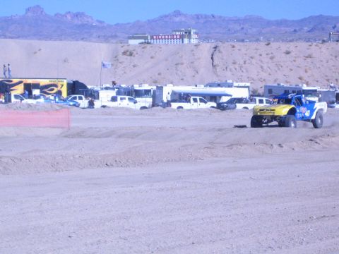 1990 Ford Bronco - yellow-and-blue off-road race truck corners on a dirt track with a Toyo Tires hauler and Riverside casino in the background.