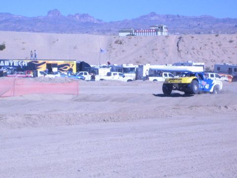 1990 Ford Bronco - yellow-and-blue No. 40 off-road race truck speeds along a dirt course past a Fabtech hauler and an orange safety fence.