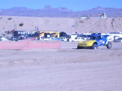 1990 Ford Bronco - yellow-and-blue No. 40 off-road race truck rounds a dirt track past a Fabtech display hauler and desert hills.