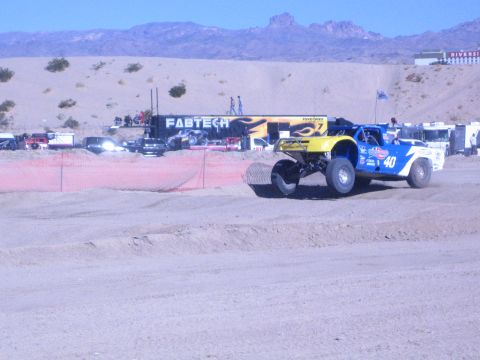 1990 Ford Bronco - yellow-and-blue No. 40 off-road race truck catches air off a dirt jump in front of a Fabtech hauler in the desert.