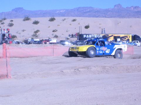 1990 Ford Bronco - yellow-and-blue No. 40 off-road race truck drives a dirt course bordered by orange safety fencing and a Fabtech hauler.