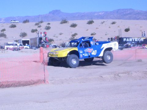 1990 Ford Bronco - blue-and-yellow No. 40 off-road race truck takes a banked dirt corner past spectators and haulers in the desert.