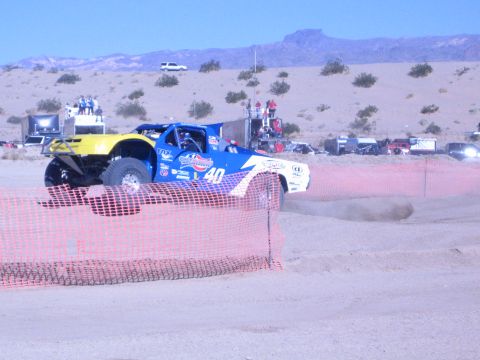 1990 Ford Bronco - blue-and-yellow No. 40 off-road race truck speeds behind an orange safety fence on a desert dirt course.