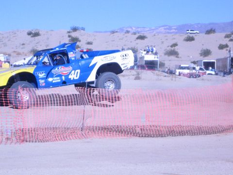 1990 Ford Bronco - blue-and-yellow No. 40 off-road race truck crests a dirt rise behind an orange safety fence in the desert.