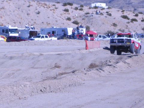 1990 Ford Bronco - red-and-white off-road race truck corners on a dirt course as spectators watch from RVs in the desert.
