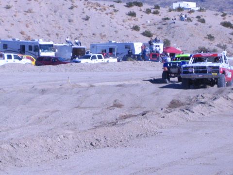 1990 Ford Bronco - Two off-road race trucks run a dirt track, one blue and one red-and-white, past RVs and pickups in the desert.